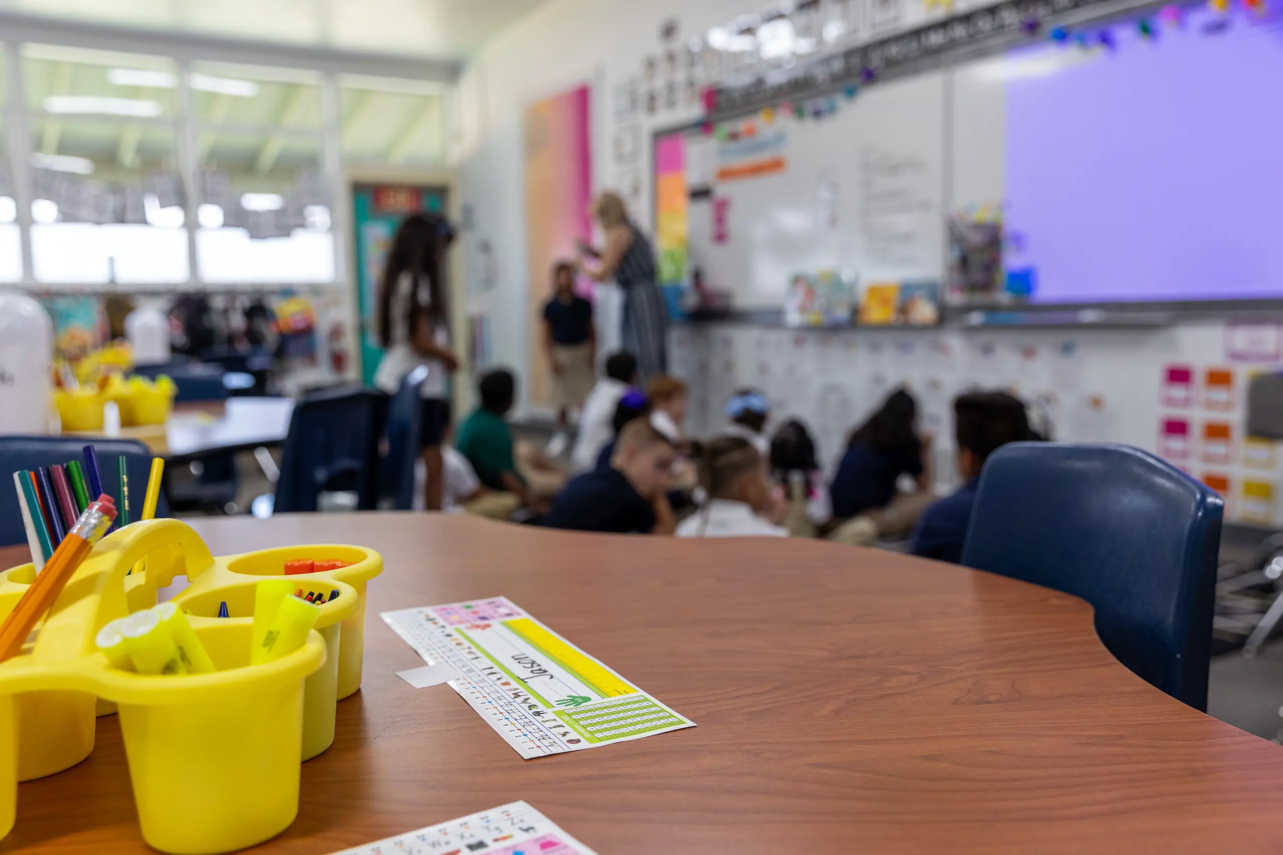 A classroom scene with students sitting on the floor near a teacher, a blurry whiteboard in the background, and a desk in the foreground with school supplies and a name tag.