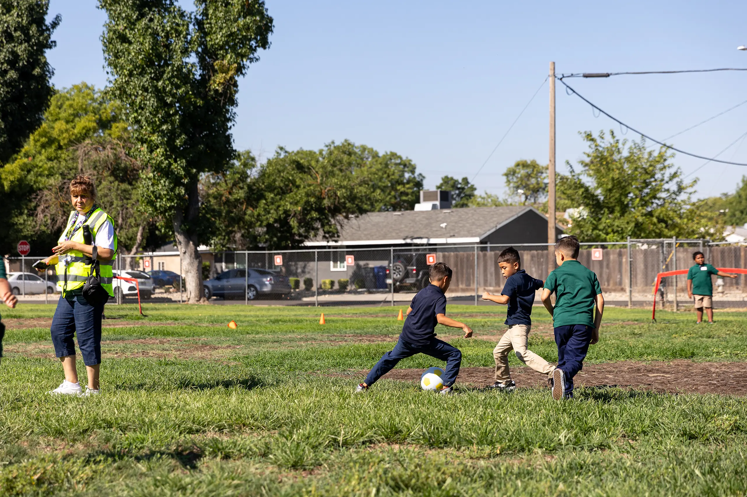 Four boys play soccer on a grassy field while an adult in a safety vest observes nearby. Trees, a fence, and houses are visible in the background under a clear sky.