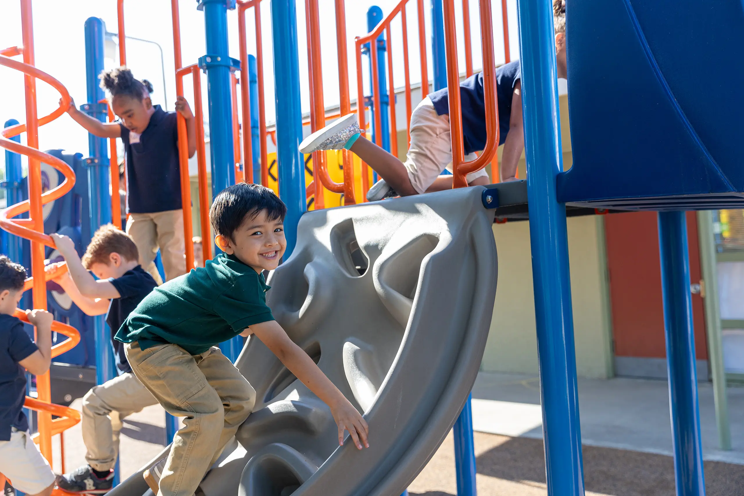 Children play on colorful playground equipment; one boy smiles as he climbs a gray climbing wall while others use orange ladders and blue bars in the background. The scene is lively and bright, suggesting a sunny day.