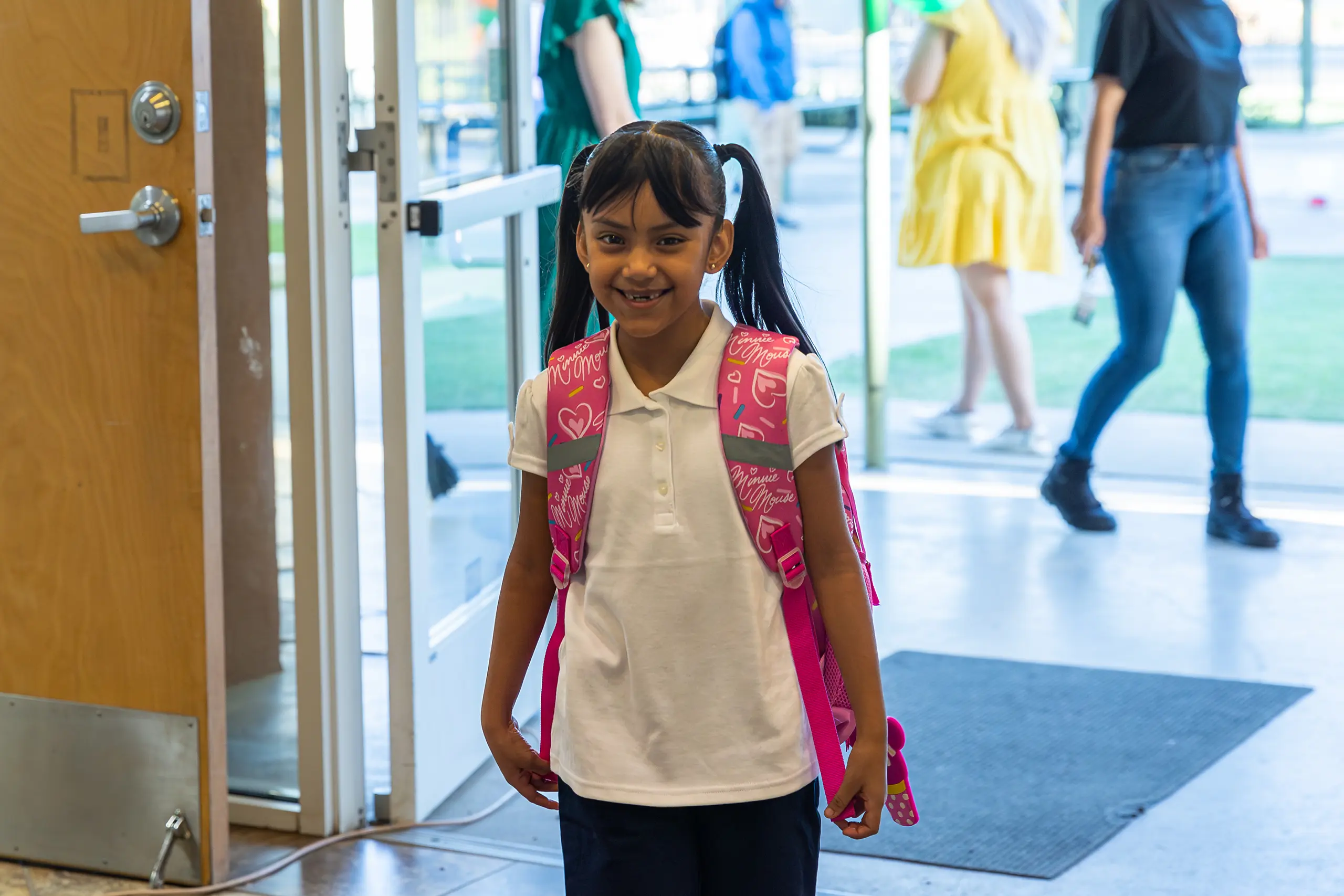 Smiling young girl with pigtails, wearing a white polo shirt and a pink backpack, stands inside near an open door with people and grass visible outside.